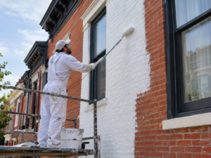 A professional painter applying a breathable mineral silicate coating to the exterior brick facade of a historic Philadelphia rowhome using a heavy nap roller.